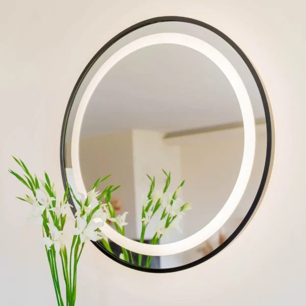 Okrągłe lustro LED w czarnej stalowej ramie loft, zawieszone na tle jasnej ściany z roślinami. Lustro loft w czarnej stalowej ramie jest dostępne w sklepie internetowym Magia Lustra – magialustra.pl.